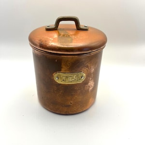 May include: A cylindrical copper tea canister with a matching lid and a brass handle. The canister features a rectangular brass plaque on the front with the word TEA embossed in the center. The copper surface shows a weathered patina.