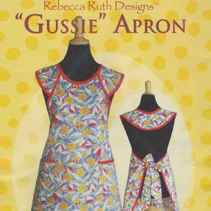 May include: A sewing pattern for the Gussie Apron by Rebecca Ruth Designs. The cover features a yellow background with white polka dots and displays two views of a floral print apron with red trim, including front and back views with a tie closure.