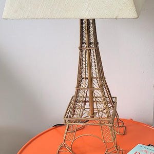 May include: A table lamp featuring a gold-colored metal base shaped like the Eiffel Tower. The lamp is topped with a square, off-white fabric lampshade with a visible woven texture. The structure is open-work metal, mimicking the tower's lattice.