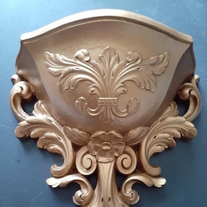 May include: A gold-colored wall-mounted planter with ornate scrollwork and a floral design. The planter is made of a lightweight material and has a curved shape.