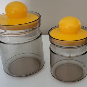 May include: Two glass jars with yellow lids and spherical knobs. The jars are clear with a smoky gray tint. The lids are a bright yellow, and the knobs are a matching yellow color. The jars are different sizes.