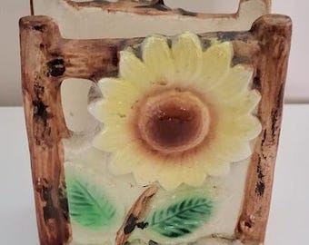 May include: A ceramic napkin holder shaped like a wooden fence with a sunflower and leaves. The sunflower is yellow with a brown center and the leaves are green.