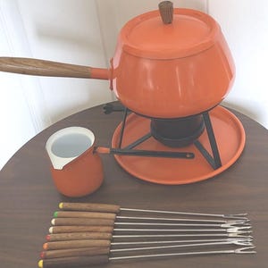 May include: An orange fondue set with a matching pot, stand, and burner. The set includes a small pitcher and several fondue forks with wooden handles and colored tips. The pot has a wooden handle and lid. The set is on a round table.