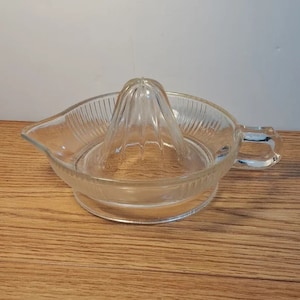 May include: A clear glass citrus juicer with a ribbed base and a cone-shaped reamer. The juicer has a handle on one side.