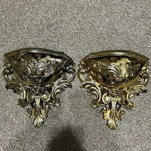 May include: Two ornate, gold-colored wall sconces with a classic, decorative design. Each sconce features a bowl-shaped top and intricate, swirling patterns. The sconces are made of a shiny, metallic material.