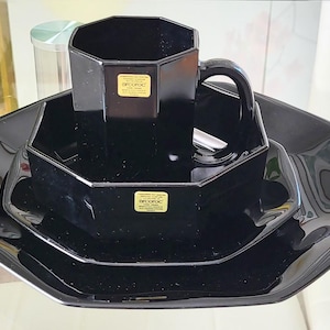 May include: A black octagonal dinnerware set, including a mug, bowl, and plates. The set has a glossy finish and a geometric design. Each piece has a small, rectangular sticker. The set is displayed on a glass surface.