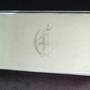 May include: A rectangular, silver-toned business card holder. The top surface features an engraved, stylized letter "G" in a decorative script. The holder has a shiny, reflective finish and a hinged lid.