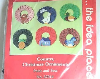 May include: A craft kit for Country Christmas Ornaments. The package features six colorful, circular ornaments with various designs, including a bear, duck, house, and pineapple. The kit is labeled "Paint and Sew" and includes the text "Mary Maxim" and "Exclusive Needlecraft Kits."