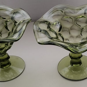 May include: Two green glass pedestal bowls with a scalloped edge and a textured surface. The bowls are perfect for serving snacks or desserts.