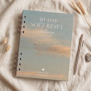 Può includere: Un quaderno a spirale con una copertina raffigurante un cielo al tramonto nei toni del blu e dell'arancione. Il testo recita 30 DAY SOFT RESET challenge e 30 days to feel like you again con una piccola icona a cuore bianco in basso.