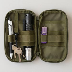 May include: An olive green zippered organizer pouch featuring internal elastic webbing and nylon straps. The left side holds a white and black pen, a black flashlight with a silver base, and two metal keys on a black clip. The right side holds a purple USB drive.