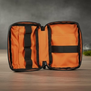 Könnte beinhalten: Ein schwarzes Reißverschluss-Etui mit leuchtend orangefarbenem Innenfutter. Das geöffnete Etui verfügt über mehrere schwarze elastische Schlaufen auf der linken Seite zur Befestigung kleiner Gegenstände und ein breites schwarzes Gummiband rechts.