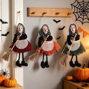 May include: Three hanging witch dolls with gray hair, black headscarves, and white aprons. Each doll wears a different colored skirt in red, orange, or green, and holds a small broomstick. They have dangling black legs and black shoes.
