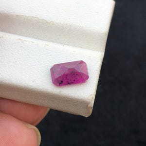 May include: A rectangular faceted gemstone with a vibrant magenta pink hue, featuring visible dark inclusions scattered throughout the stone's surface, showcasing a clear, polished finish with sharp, defined edges and a flat top surface.