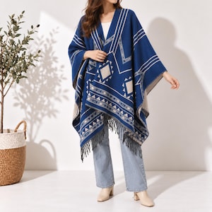 May include: A navy blue poncho featuring a cream-colored geometric pattern with diamonds and triangles. The garment has an open front, wide sleeves, and a hemline finished with black fringe. It is worn over a white top and light blue denim jeans.