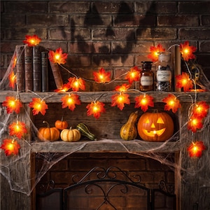 May include: A string of illuminated maple leaf lights draped across a wooden mantelpiece. The orange and red leaves glow with warm light, interspersed with small pumpkins, a carved jack-o'-lantern, old books, and an apothecary bottle.