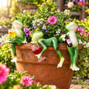 May include: Three green frog figurines perched on the rim of a large terracotta pot. One frog holds a yellow drink, another reclines while holding a glass of red wine, and the third stands upright holding a small bottle of red wine.