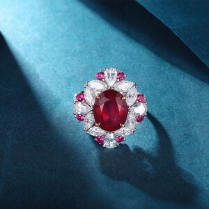 Può includere: Un anello con un grande rubino rosso intenso a taglio ovale al centro, circondato da un gruppo di pietre bianche trasparenti a taglio marquise e pietre rosse più piccole a taglio rotondo su una fascia metallica, creando un motivo floreale.