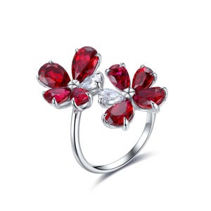 May include: A silver-toned open ring featuring two flower-shaped clusters. Each cluster is composed of multiple pear-cut deep red stones and a single pear-cut clear white stone, all set in a polished metal band with a prong setting design.