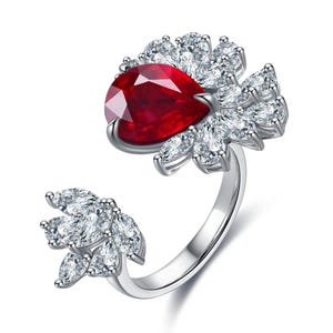 Op de afbeelding: Een open zilveren ring met een grote peervormige dieprode edelsteen als middelpunt, omringd door een cluster van helderwitte marquise-geslepen stenen. Een kleiner bijpassend cluster van witte marquise-geslepen stenen bevindt zich aan het andere uiteinde.