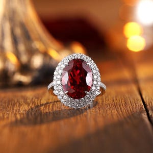 Op de afbeelding: Een ring met een grote, ovaal geslepen dieprode edelsteen in het midden, omringd door een dubbele halo van kleine, heldere, rond geslepen stenen in een metalen band. Het ontwerp bevat klauwzettingen en een pavé-zetting voor de stenen.