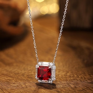May include: A silver-toned necklace featuring a square-cut deep red gemstone pendant. The center stone is surrounded by a halo of smaller, clear baguette and round-cut stones, suspended from a delicate, thin silver-toned metal chain.