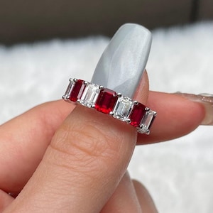 Pode incluir: Um anel de eternidade em tom prateado com um padrão alternado de rubis vermelho-escuro e diamantes brancos transparentes de corte retangular. As pedras estão cravadas numa montagem de garras partilhadas que rodeia todo o aro do anel.