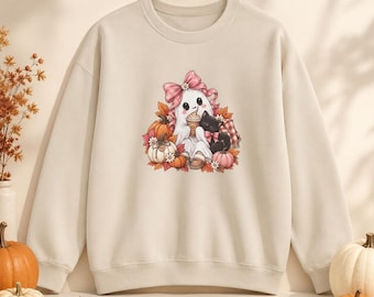 Könnte beinhalten: Ein beigefarbenes Sweatshirt mit Rundhalsausschnitt und einem zentralen Aufdruck eines weißen Geistes mit großer rosa Schleife, der einen Kaffeebecher hält und mit einer schwarzen Katze zwischen orangefarbenen Kürbissen sitzt.