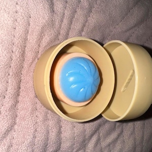 Peut inclure: Un récipient en plastique beige avec un couvercle amovible, contenant un objet circulaire avec une base couleur pêche et un centre bleu surélevé en forme de spirale. Le récipient est cylindrique avec des parois lisses et un design simple.