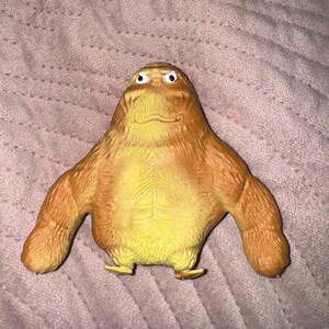 May include: A brown, muscular, ape-like creature figurine with a textured, fur-like surface, defined abdominal muscles, and small white eyes with black pupils. The figure has a wide, rounded head, broad shoulders, and two long, thick arms.