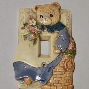 May include: A decorative light switch cover featuring a tan teddy bear with a blue scarf and outfit. The bear is holding a bouquet of pink flowers. The cover has a cream-colored background with a basket and floral accents.