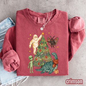 May include: A brick red crewneck sweatshirt featuring a graphic of a yeti, a moth, an alien, and a sea creature decorating a Christmas tree. The inner neck label reads Comfort Colors. A silver chain necklace rests on the collar of the garment.