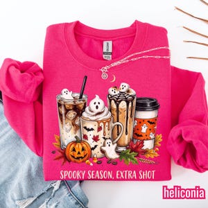Puede incluir: Una sudadera de manga larga en color rosa brillante con un gráfico de cuatro bebidas de café, una calabaza y fantasmas. El texto dice SPOOKY SEASON, EXTRA SHOT. El diseño incluye hojas de otoño, murciélagos y malvaviscos. Un collar de plata descansa en el cuello.
