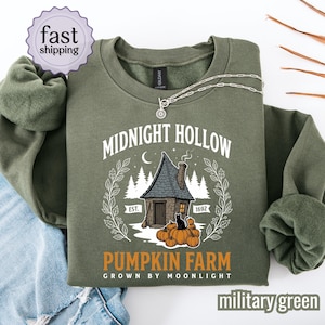 May include: A military green crewneck sweatshirt featuring a graphic of a house, pumpkins, and a black cat. The text reads Midnight Hollow Pumpkin Farm Est. 1692 Grown by Moonlight. A silver chain necklace rests on the collar of the garment.