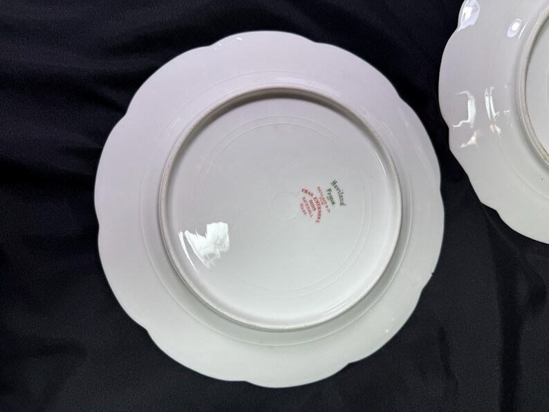May include: Two white porcelain plates with scalloped edges. The underside of one plate shows the text "Haviland France" in red lettering. The plates are on a dark surface.