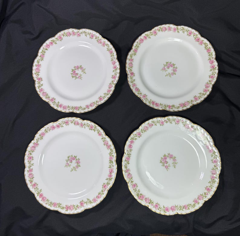 May include: Four white plates with scalloped edges, each adorned with a delicate pink floral border and a central floral cluster. The plates have a gold trim.