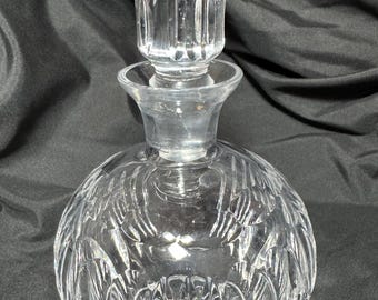 May include: A clear, round crystal decanter with a matching stopper. The decanter has a detailed, etched pattern on the body and a ribbed stopper. The glass is transparent, allowing light to pass through. A decorative piece for serving beverages.