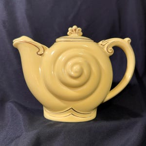 Peut inclure: Une théière en céramique jaune pâle présentant un motif en spirale rappelant un coquillage sur son corps. La théière comprend un couvercle assorti avec un petit bouton décoratif, une anse courbée et un bec verseur, soulignés de doré.