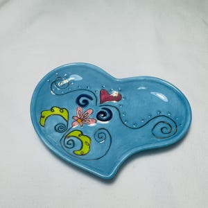 May include: A heart-shaped ceramic dish in a light blue hue. The dish is decorated with a pink heart, a pink and white flower, and green and yellow leaf-like shapes. Black swirls and dots add detail.