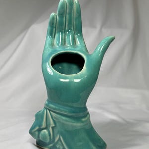 May include: A turquoise ceramic hand-shaped vase with a central opening. The vase features a detailed design of a hand with fingers extended and a decorative base. The glossy finish enhances the vibrant color, making it a unique decorative item.