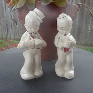 May include: Two vintage ceramic figurines of boys in white outfits, one with a top hat and the other with a cap. Both are playing musical instruments with red accents. The figurines are standing on a dark surface.