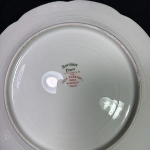May include: White porcelain plate with a scalloped edge. The underside features the text "Haviland France" and "CHAS. EMERSON SONS HAVERHILL MASS." in red lettering. The plate has a glossy finish.