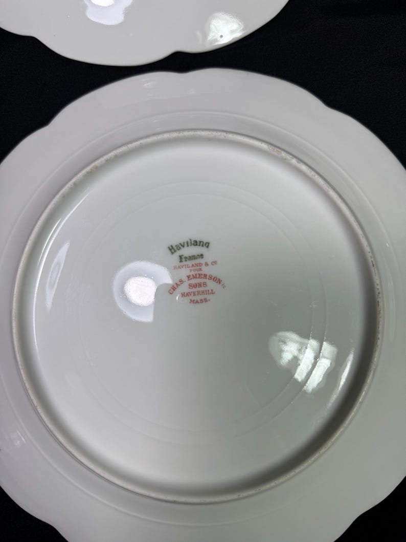May include: White porcelain plate with a scalloped edge. The underside features the text "Haviland France" and "CHAS. EMERSON SONS HAVERHILL MASS." in red lettering. The plate has a glossy finish.
