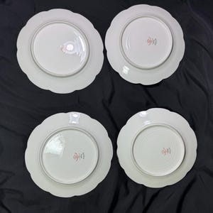 May include: Four white ceramic plates with scalloped edges are arranged on a black surface. Each plate has a central, slightly recessed area. Red Japanese characters are printed on the center of each plate.