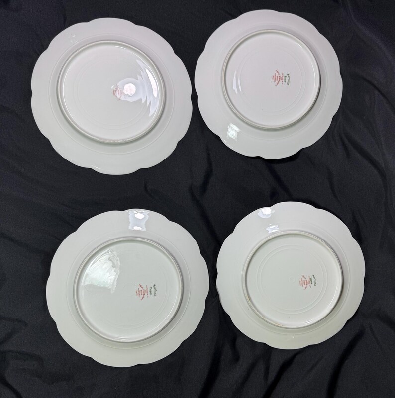 May include: Four white ceramic plates with scalloped edges are arranged on a black surface. Each plate has a central, slightly recessed area. Red Japanese characters are printed on the center of each plate.