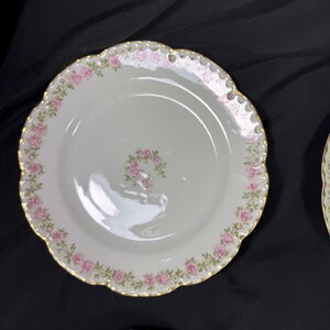 May include: White porcelain plate with a scalloped edge and gold trim. The plate is decorated with a border of pink roses and a small cluster of roses in the center. The plate is likely part of a vintage dinnerware set.