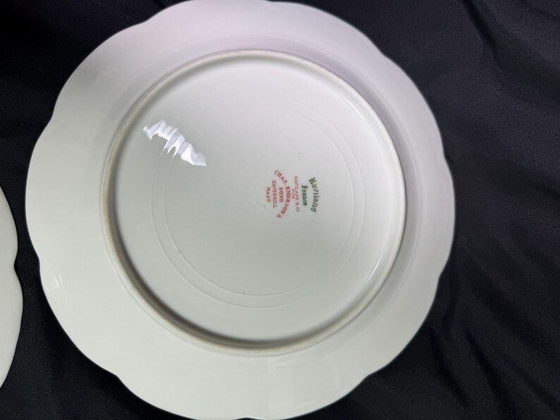 May include: White porcelain plate with a scalloped edge. The underside features a circular design and text in red and green, including "Haviland France" and "Limoges". The plate has a glossy finish and appears to be vintage.