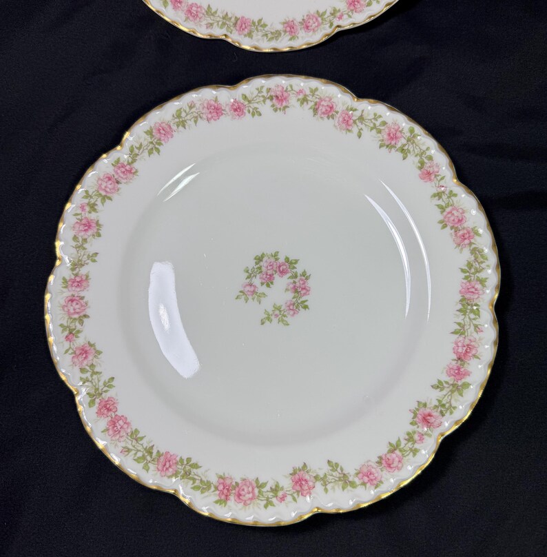 May include: White porcelain plates with a scalloped edge and a gold trim. The plates are decorated with a pink rose garland around the rim and a small rose wreath in the center. The plates are on a dark background.