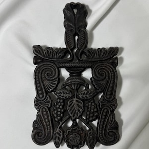 May include: A rectangular black cast iron trivet featuring an intricate openwork design. The pattern includes symmetrical scrollwork along the sides, a central motif of leaves and berries, and a decorative handle at the top with leaf detailing.