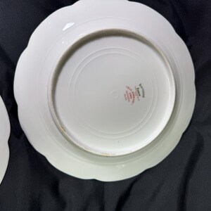 May include: Two white ceramic plates with scalloped edges. The plates have a circular design in the center and a red stamp with text. The plates are on a black surface.